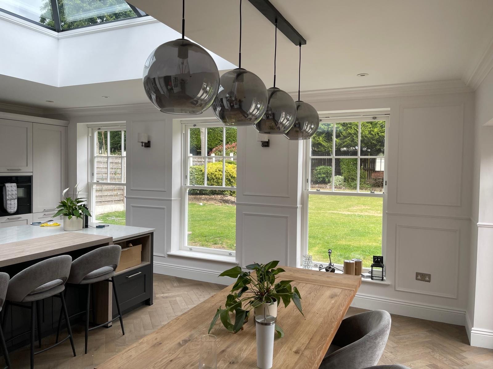 Grey Shaker kitchen island with smoked glass pendant lights, dining table and windows onto the garden